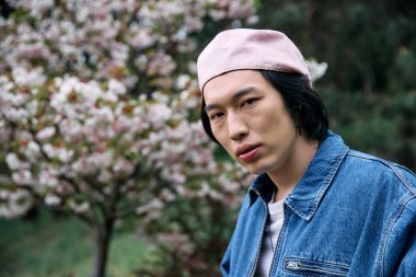 Genç Asyalı bir adam, bir parkta çiçeklerin açtığı şık bir kot elbiseyle güvenle poz veriyor..