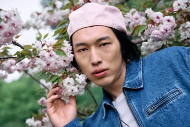 Genç Asyalı adam, bahar aylarında canlı kiraz çiçekleri arasında kot elbisesiyle rahatça poz veriyor..