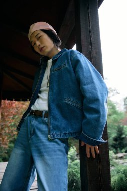 Genç Asyalı bir adam lüks bir park ortamında rahatlarken şık kot kıyafetini gösteriyor..