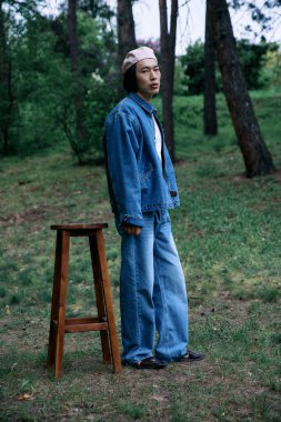 Huzurlu bir parkta, genç bir adam, doğa ile çevrili, modaya uygun bir kotla güvenle ayakta duruyor..