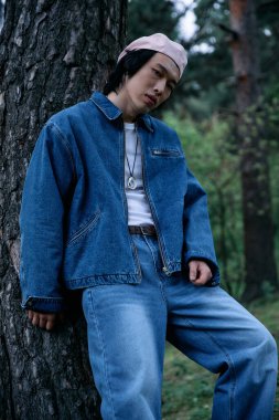 Yeşillik bir parkta, genç bir adam ağaçta dinlenirken modern kot tarzını sergiliyor..