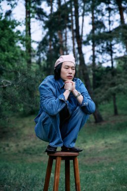 Genç bir Asyalı parkta bir taburede oturuyor, şık bir kot giysisi ve neşeli bir pozu var..