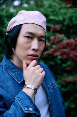 Genç Asyalı adam, parkta yeşilliklerle çevrili bir kot elbiseyle güven içinde poz veriyor..