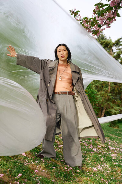 Молодой азиат в сером плаще стоит в парке, позирует рядом с полиэтиленом, концепция устойчивости