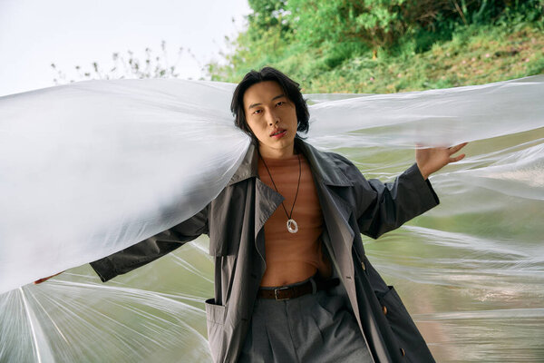В оживленном парке молодой азиат моделирует модный серый плащ, позирует рядом с полиэтиленом, концепция устойчивости