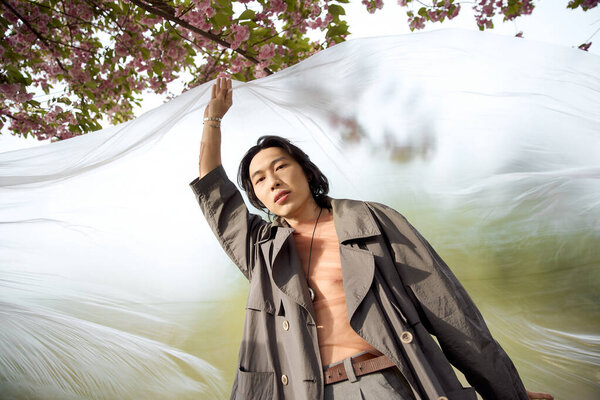 Молодой азиат позирует в парке, демонстрируя современную моду среди цветущей вишни.