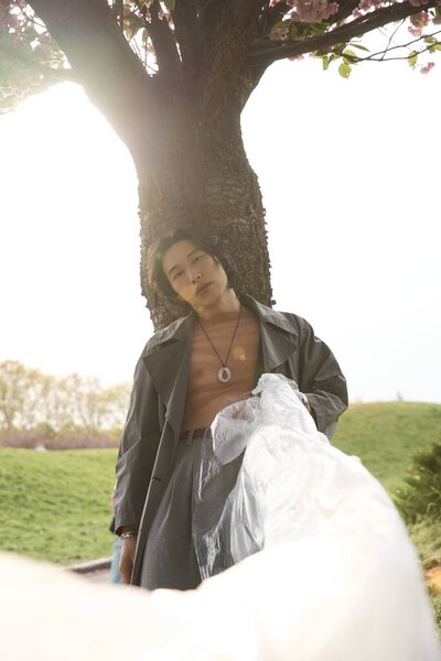 В мирном парке молодой человек стоит стильно задрапированный в серый, с полиэтиленом, обнимающий теплый весенний свет.