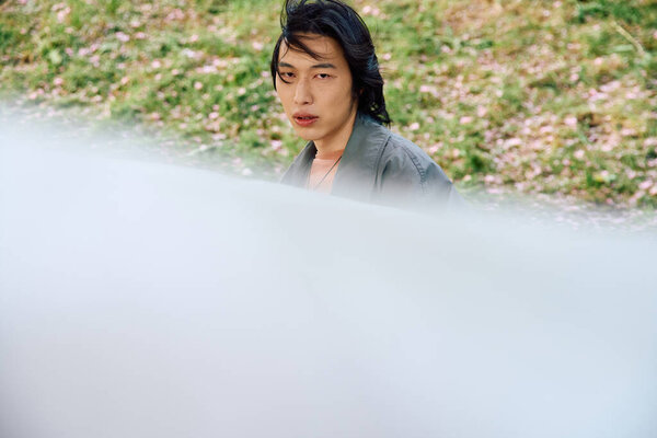 A young Asian man strolls through a peaceful park wearing a trendy grey outfit.