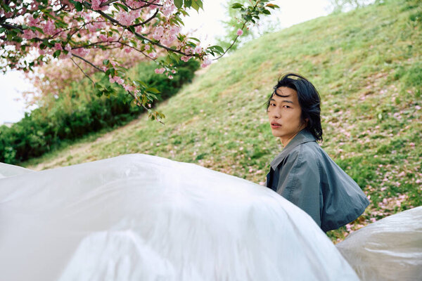 Молодой азиат в стильном сером наряде позирует с полиэтиленом рядом с цветущими цветами в парке.
