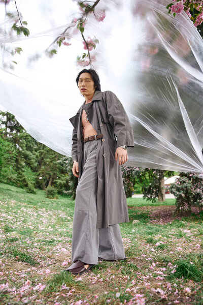 Молодой азиат демонстрирует свой модный серый наряд, гуляя рядом с полиэтиленом в живописном парке.