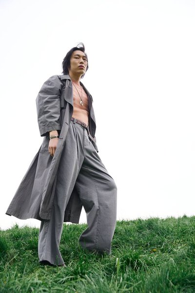 Молодой азиат моделирует свой стильный серый наряд на зеленой траве, наслаждаясь природой.