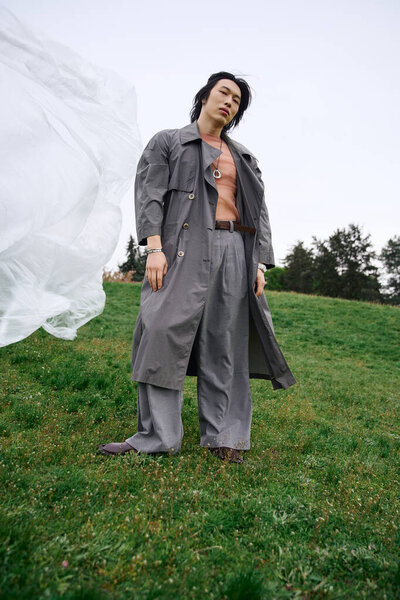Молодой азиат уверенно стоит в сером плаще посреди зеленой травы в парке.