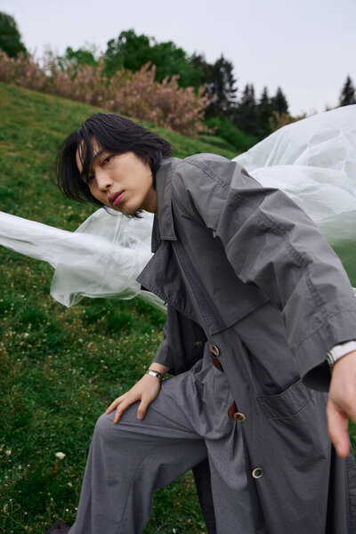 Молодой азиат демонстрирует современную моду в стильном сером плаще возле полиэтилена на траве