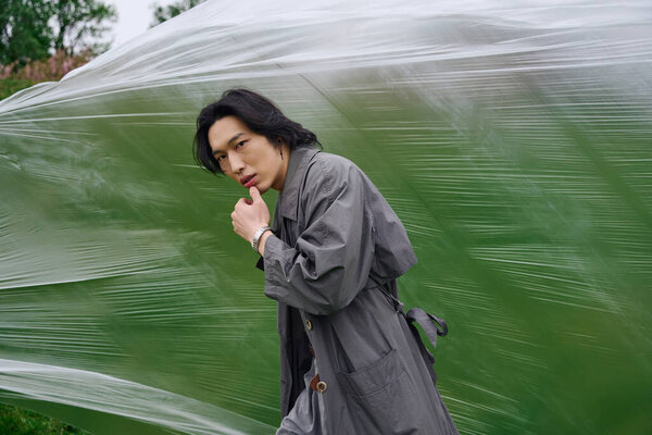 A young asian man strolls through the park, exuding confidence in a fashionable grey outfit.