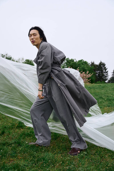 Молодой человек, одетый в модный серый плащ, позирующий возле полиэтилена на траве