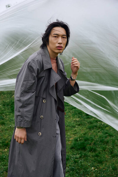 Молодой азиат в модном сером плаще стоит рядом с полиэтиленом среди пышной зелени, привлекая внимание.