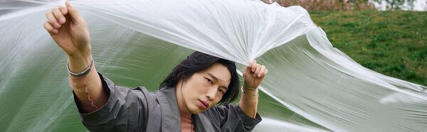 Молодой азиат моделирует модный серый плащ рядом с полиэтиленом, баннер