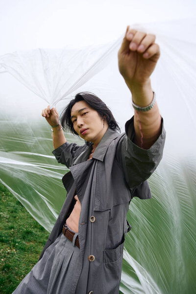 Молодой азиат в сером плаще уверенно стоит среди пышной зелени, держа в руках пластик