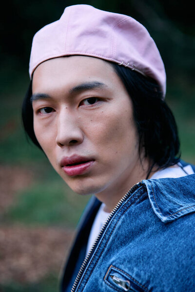 A young asian man showcases a chic denim look while relaxing in a vibrant park setting.