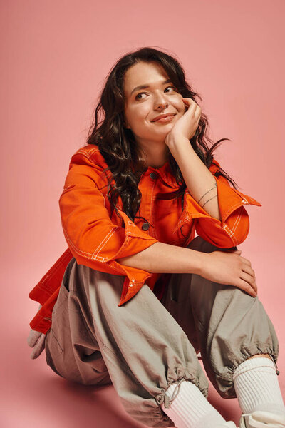 Stylish young woman poses thoughtfully while wearing bright attire against pink background.