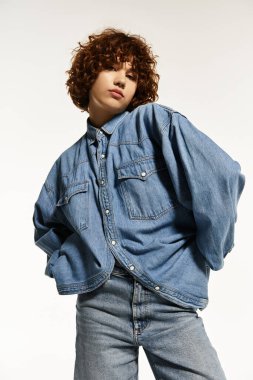 Genç bir kadın kot gömlek ve kot pantolon içinde beyaz bir fona karşı güvenle poz veriyor..