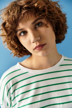 Kıvırcık saçlı genç bir kadın canlı bir elbise içinde mavi bir fona karşı poz veriyor..