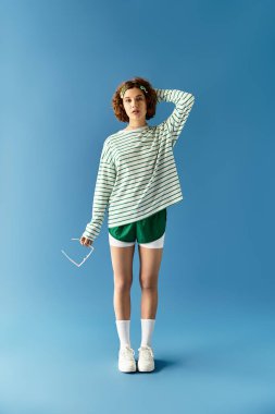 Genç bir kadın parlak mavi bir arka plana karşı şık yaz kıyafetiyle hava atıyor, neşe saçıyor.