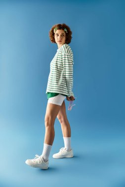 Kısa kıvırcık saçlı genç bir kadın canlı yaz tarzını mavi bir arka planda sergiliyor..