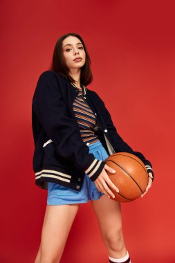 Modaya uygun giyinmiş genç bir kadın dikkat çekici bir ortamda kendine güvenerek basketbol topuyla poz veriyor..