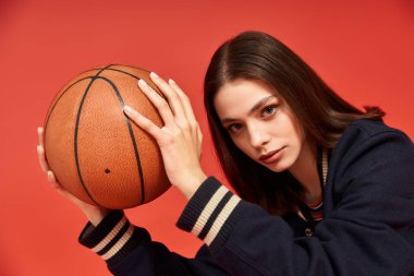 Moda öncüsü kadın canlı kırmızı bir arka plana karşı kendine güvenen bir basketbol topuyla poz veriyor..