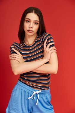 Renkli giyinmiş genç bir kadın dikkat çekici bir kırmızı arka planla güvenle ayakta duruyor.