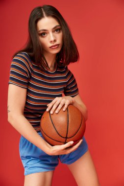 Modaya uygun genç bir kadın elinde basketbol topuyla dikkat çekici bir kırmızı zemine karşı duruyor..