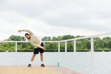 Kapüşonlu formalı genç adam sakin suyun yanında esneme egzersizi yapıyor, doğayı kucaklıyor..