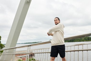 Kapüşonlu genç adam bulutlu bir gökyüzünün altında sakin bir nehre bakan bir köprüye uzanır..