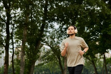 Sakallı genç bir adam yeşil bir parkta koşar, formda kalır ve egzersiz rutinine odaklanır..