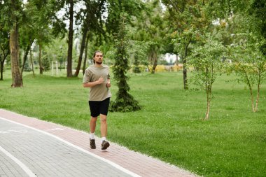 Sakallı genç bir adam yemyeşil bir parkta koşuyor, vücut bulmuş zindelik ve canlılık..