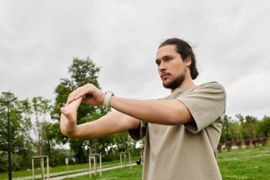 Sakallı genç bir adam dışarıda parkta egzersiz yaparken kollarını uzatıyor..