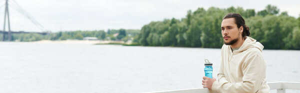 A young sporty man in a hoodie takes a moment to relax by the water while exercising outdoors, banner