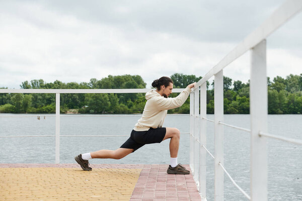 A young man in a hoodie lunges on a dock, relishing the fresh air and calm waters.