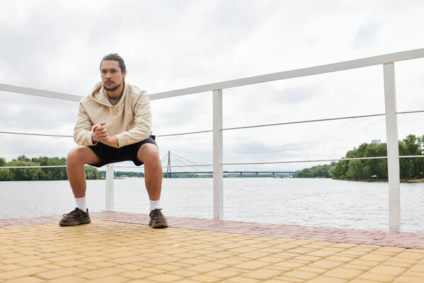 Young man exercises in a hoodie outdoors, focused and determined beside the riverbank.