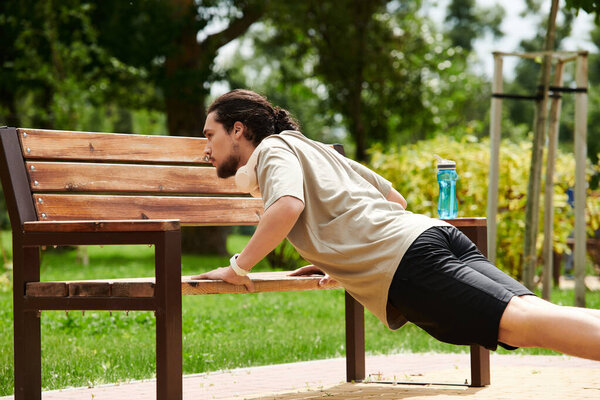 A fit young man with a beard enjoys exercising outdoors by performing push-ups on a bench.