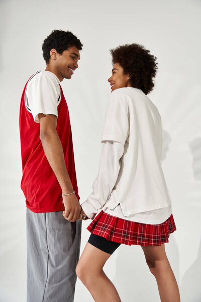 Two young lovers in trendy clothes hold hands and smile, radiating affection.