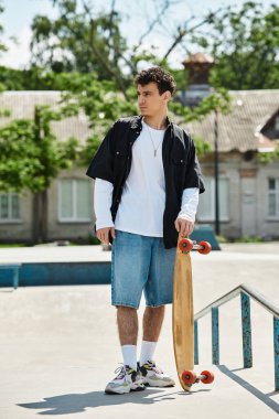 Genç bir adam parkta dikiliyor, üstünkörü giyinmiş ve kaykayını tutuyor..