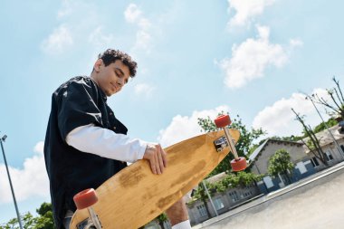 Genç bir adam parkta elinde uzun tahtasıyla duruyor ve önündeki güzel günün tadını çıkarıyor..