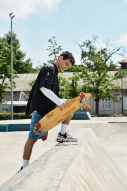 Genç bir adam parktaki kaykay rampasında duruyor, açık gökyüzünün altında kaykaya binmeye hazırlanıyor..