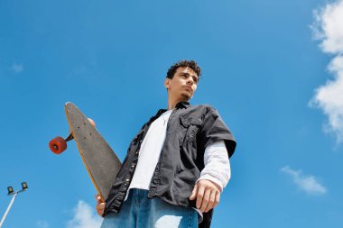 Kaykaycı açık havada eğleniyor, kendine güvenen bir kaykay tutuyor ve ufka bakıyor.