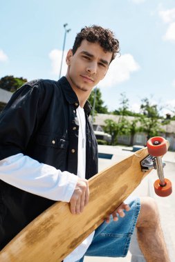 Genç bir adam kaykayıyla parkta oturuyor, parlak gökyüzüne karşı güvenle poz veriyor..