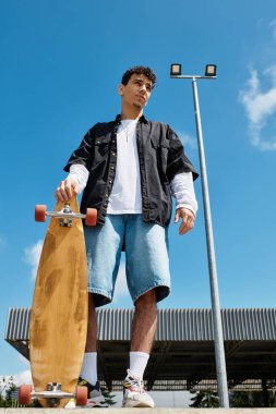 Genç bir adam güvenle parkta duruyor, yanında kaykay, açık mavi bir gökyüzünün altında..