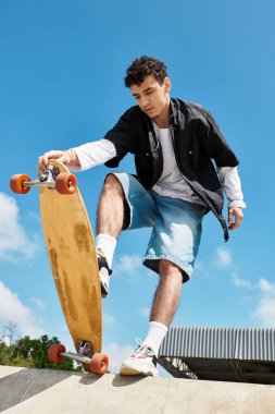 Genç bir adam parlak mavi gökyüzünün altındaki canlı bir parkta kaykay yeteneğini sergiliyor..