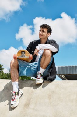 Genç bir adam beton bir kenarda oturuyor, elinde kaykayla güneşli park atmosferinin tadını çıkarıyor..